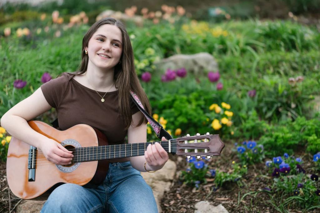 Kellis's senior photos taken Downtown, Greenville, SC: Young woman sitting outdoors in a garden, playing an acoustic guitar and smiling, surrounded by colorful flowers and greenery.