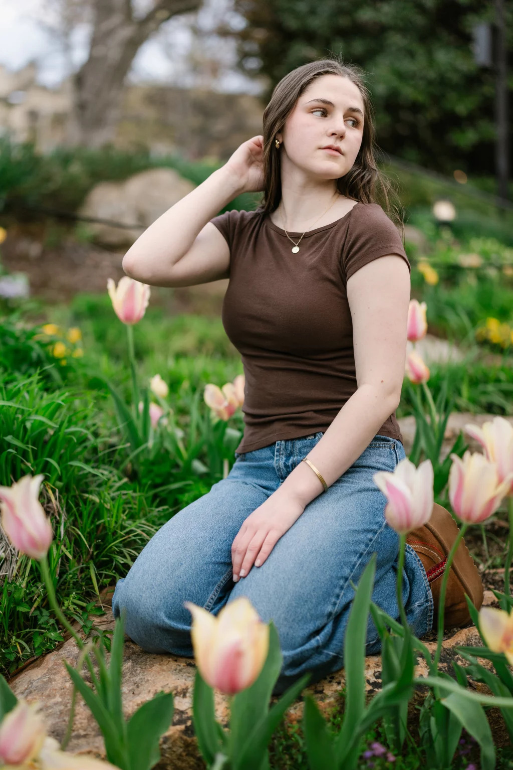 Kellis's senior photos taken Downtown, Greenville, SC: A young woman with long brown hair, wearing a brown shirt and blue jeans, kneels on a rock in a garden surrounded by pink and yellow tulips, looking to the side with one hand touching her hair.