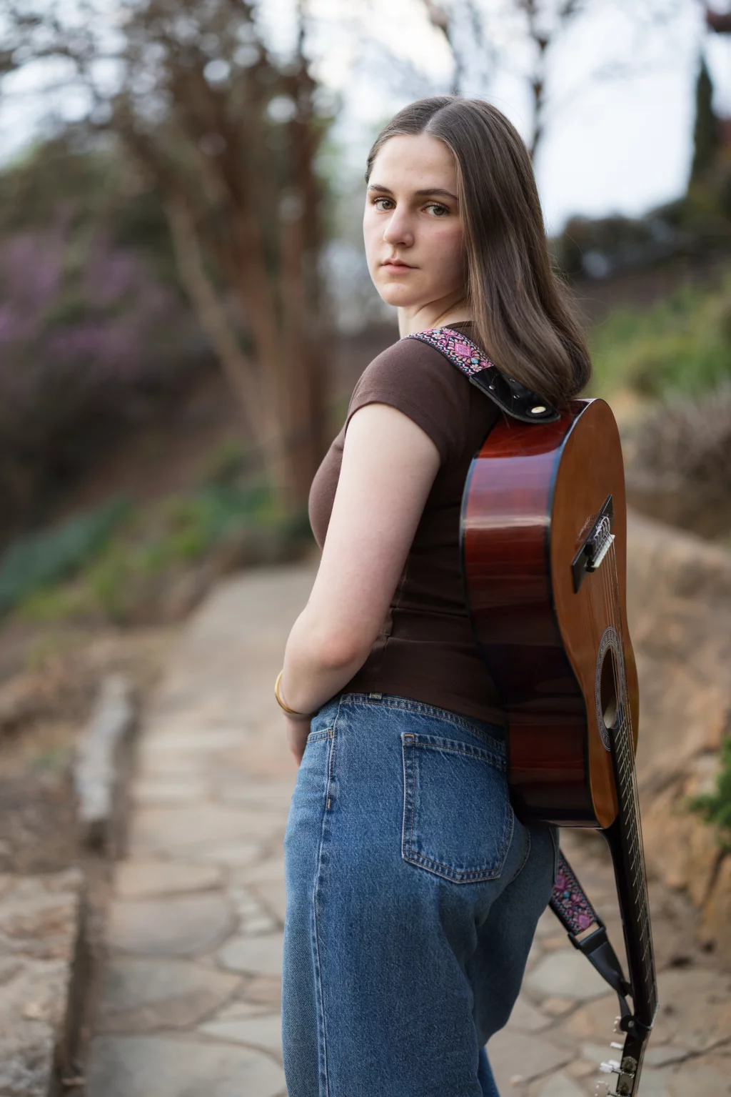 Kellis's senior photos taken Downtown, Greenville, SC: A young woman with straight brown hair stands outdoors on a stone path, looking over her shoulder at the camera. She is wearing a brown t-shirt, jeans, and has an acoustic guitar strapped across her back. The background is blurred with trees and greenery.