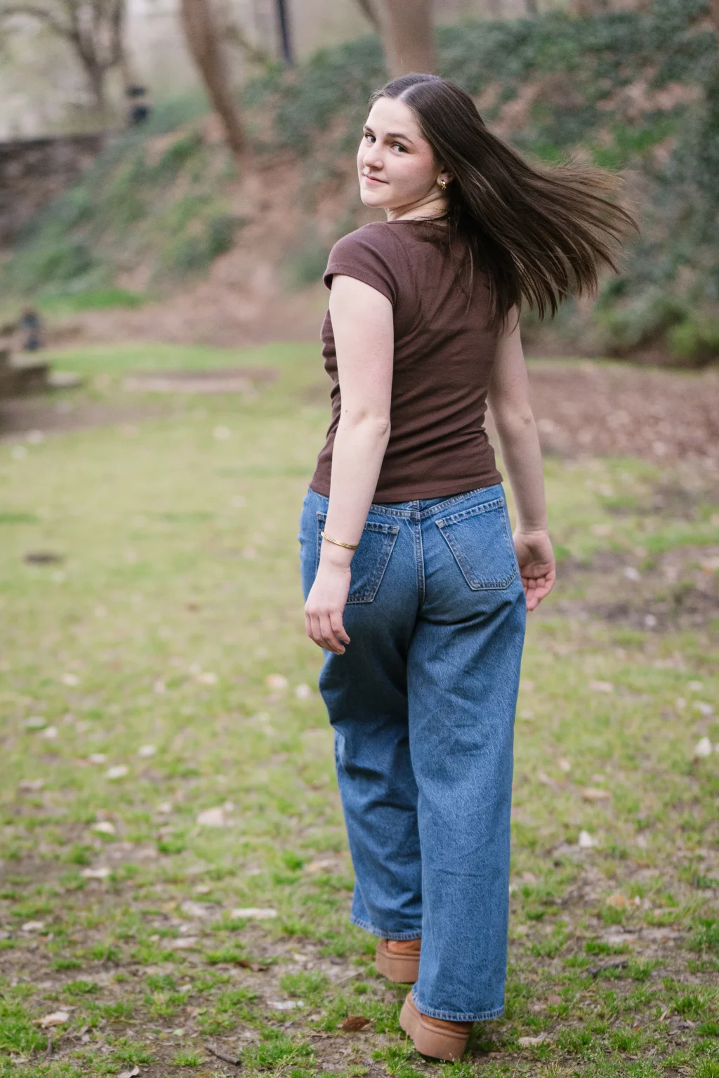 Kellis's senior photos taken Downtown, Greenville, SC: Young woman with long brown hair, wearing a brown t-shirt and blue jeans, walking outdoors on grass and looking back over her shoulder.