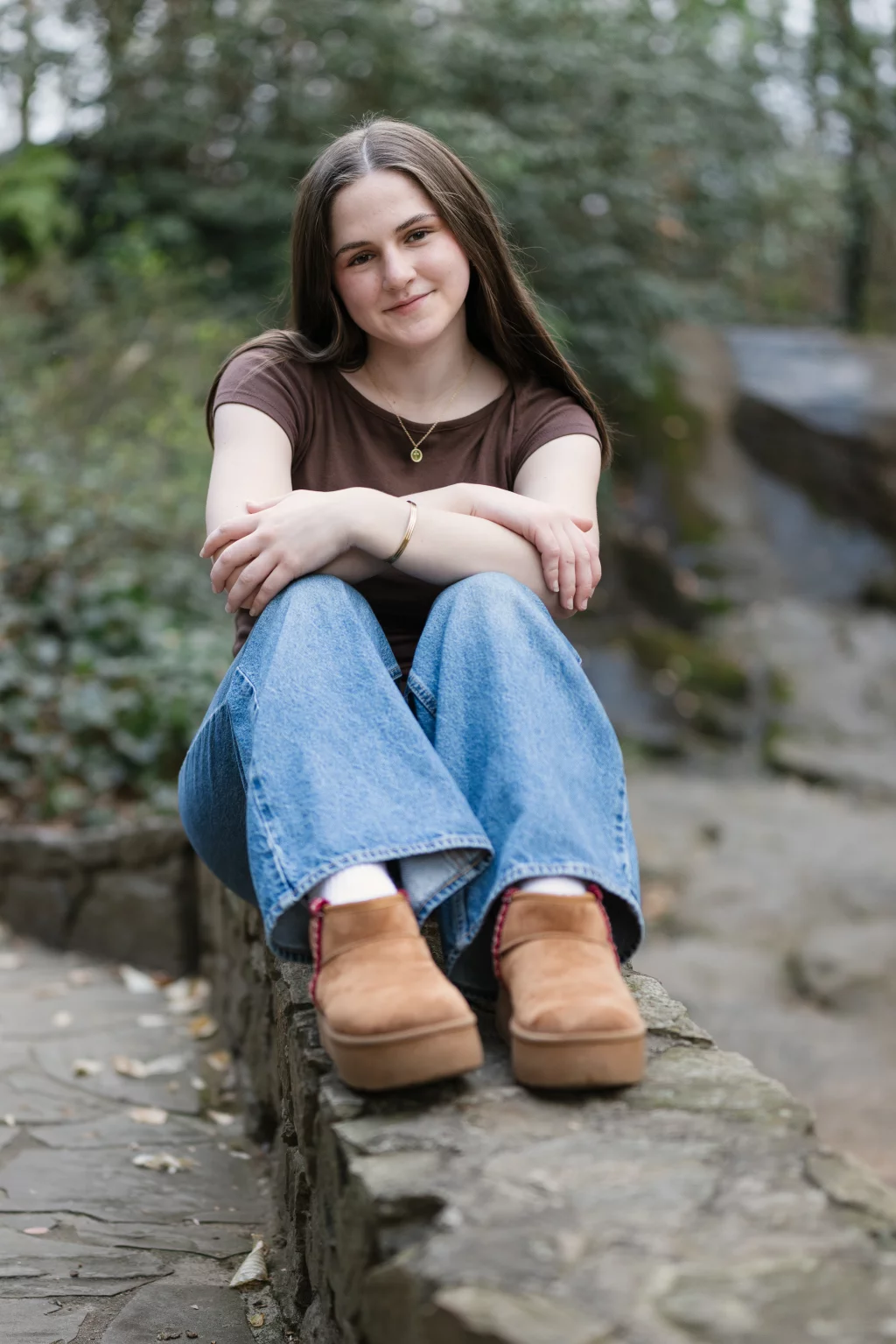 Kellis's senior photos taken Downtown, Greenville, SC: A young woman with long brown hair, wearing a brown shirt, blue jeans, and tan shoes, sits on a stone ledge outdoors with her arms wrapped around her knees and a slight smile on her face. The background is blurred greenery.