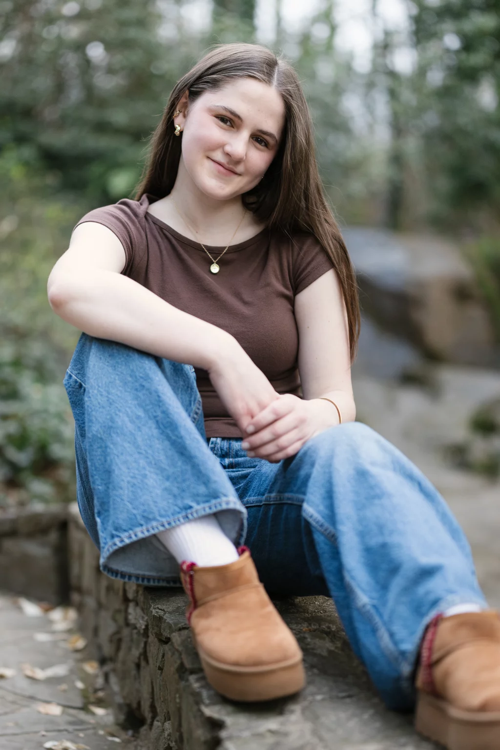 Kellis's senior photos taken Downtown, Greenville, SC: Young woman with long brown hair sitting outdoors on a stone ledge, wearing a brown t-shirt, blue jeans, white socks, and tan slip-on shoes, smiling at the camera with greenery in the background.