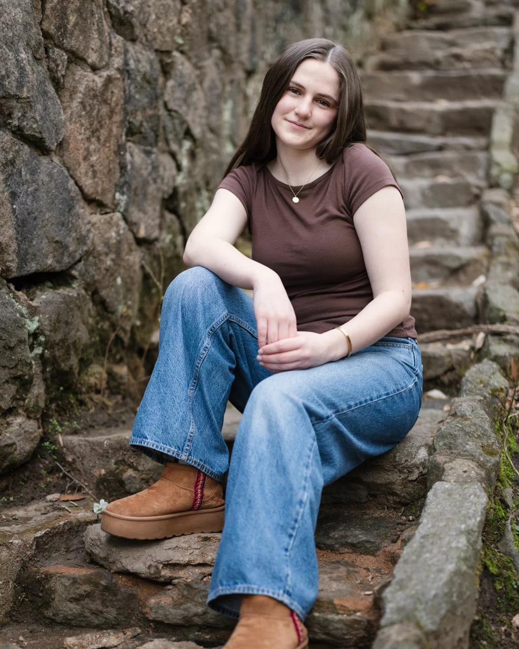 Kellis's senior photos taken Downtown, Greenville, SC: Young woman with long brown hair sits on outdoor stone steps, wearing a brown t-shirt, blue jeans, and tan boots, with a stone wall beside her.