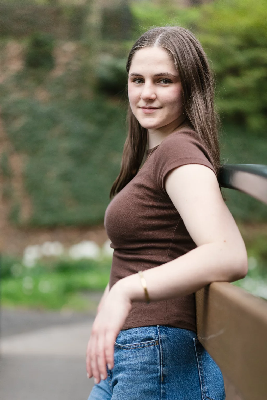 Kellis's senior photos taken Downtown, Greenville, SC: Young woman with long brown hair wearing a brown t-shirt and blue jeans, sitting on a bench outdoors and looking at the camera, with a blurred green background.