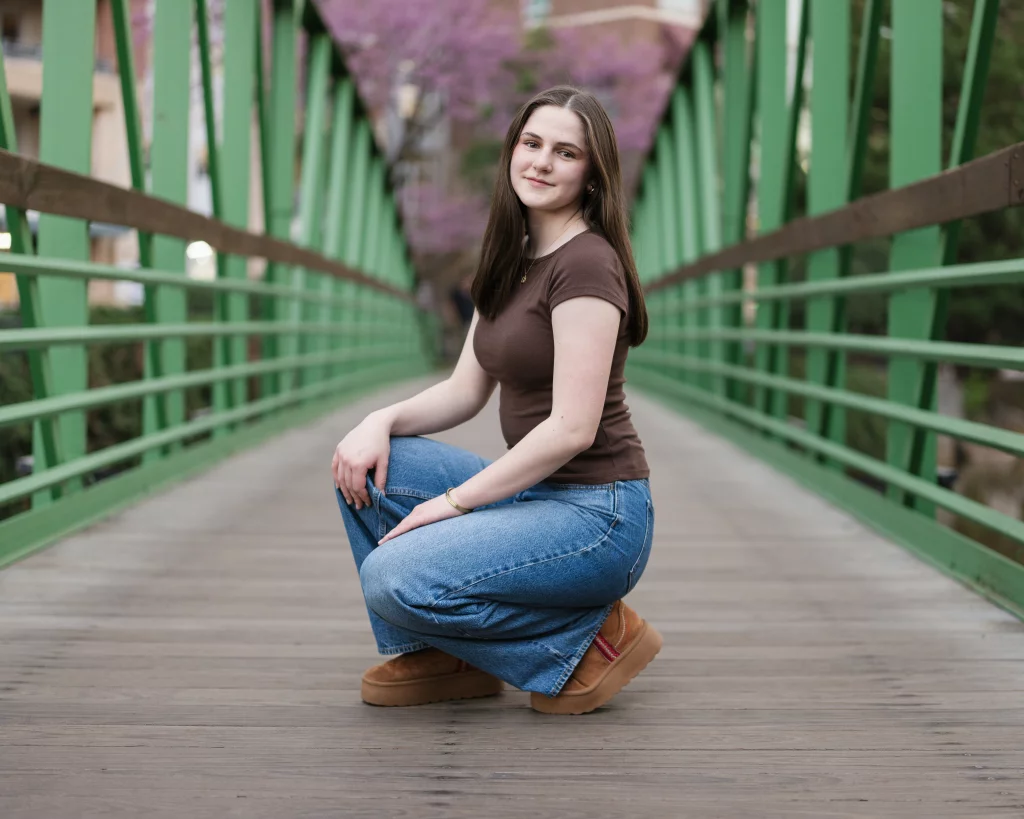 Kellis's senior photos taken Downtown, Greenville, SC: Young woman with long brown hair, wearing a brown shirt and blue jeans, squatting on a green metal bridge with wooden planks and pink-blossomed trees in the background.