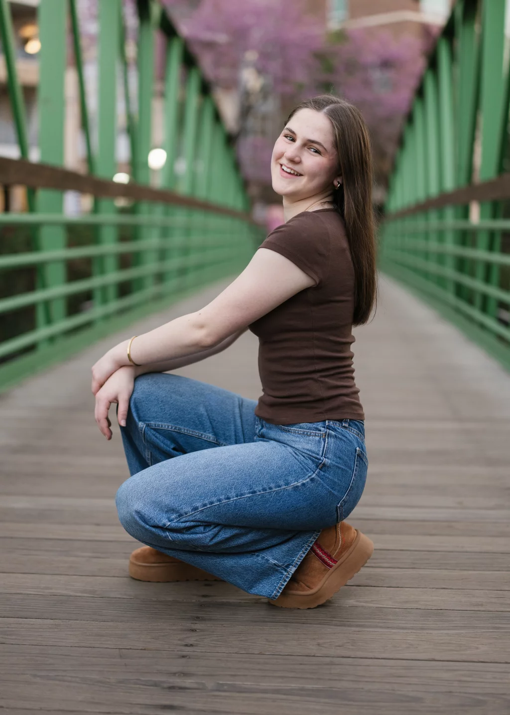 Kellis's senior photos taken Downtown, Greenville, SC: A young woman with long brown hair kneels and smiles on a green pedestrian bridge with blooming trees in the background. She is wearing a brown shirt, blue jeans, and tan shoes.
