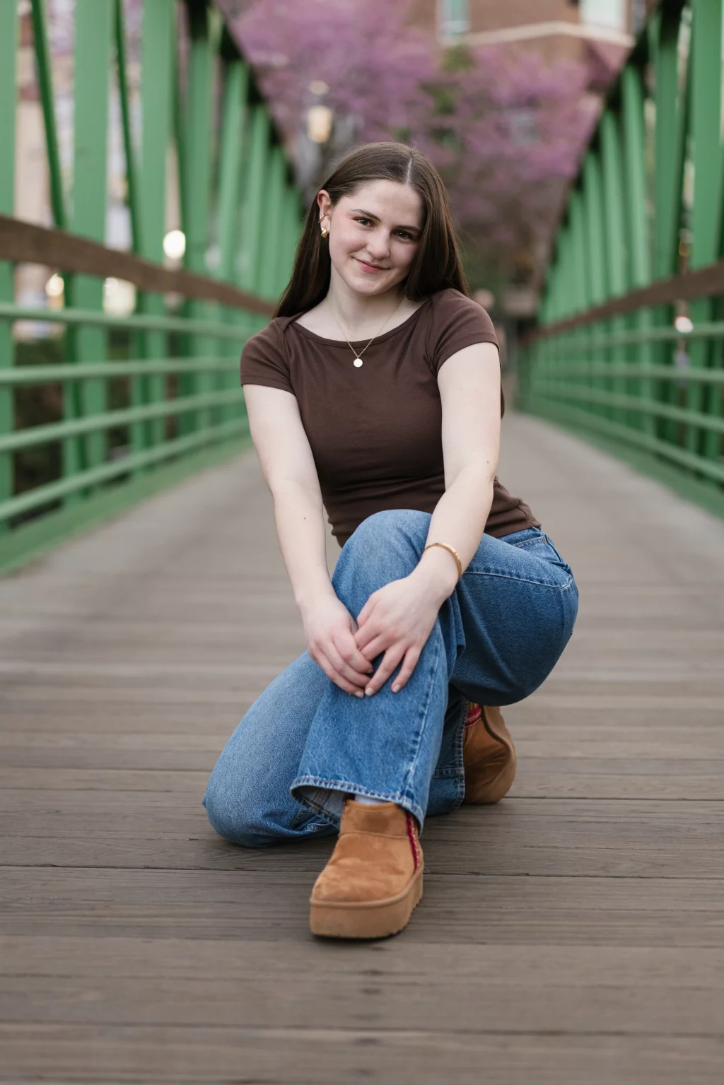 Kellis's senior photos taken Downtown, Greenville, SC: Young woman with long brown hair, wearing a brown t-shirt, blue jeans, and tan platform shoes, kneeling and smiling on a wooden pedestrian bridge with green railings and blurred pink blooming trees in the background.