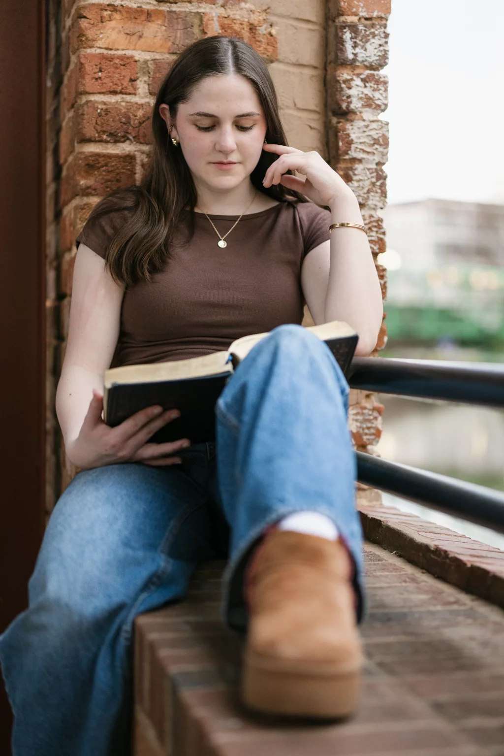 Kellis's senior photos taken Downtown, Greenville, SC: A young woman with long brown hair sits on a brick ledge, reading a book. She is wearing a brown shirt, blue jeans, and tan shoes, with one leg bent and resting on the ledge.