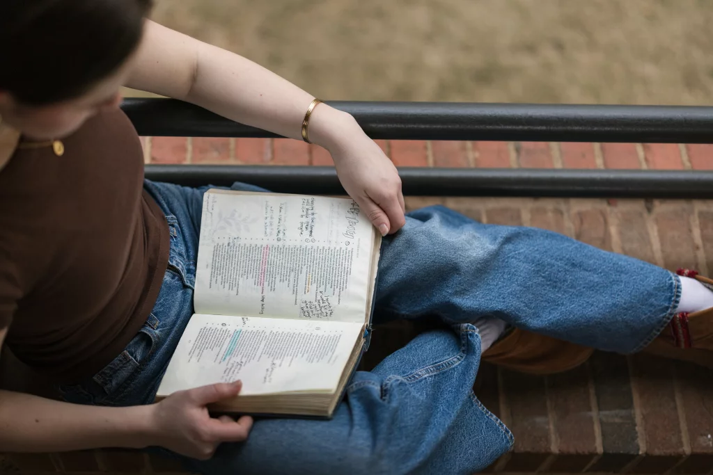 Kellis's senior photos taken Downtown, Greenville, SC: Person sitting on a brick ledge, wearing jeans and a brown shirt, reading an open book with handwritten notes in the margins.