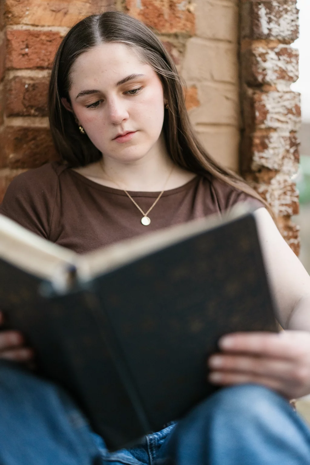 Kellis's senior photos taken Downtown, Greenville, SC: Young woman with long brown hair, wearing a brown shirt and gold necklace, sitting against a brick wall and reading a large book.