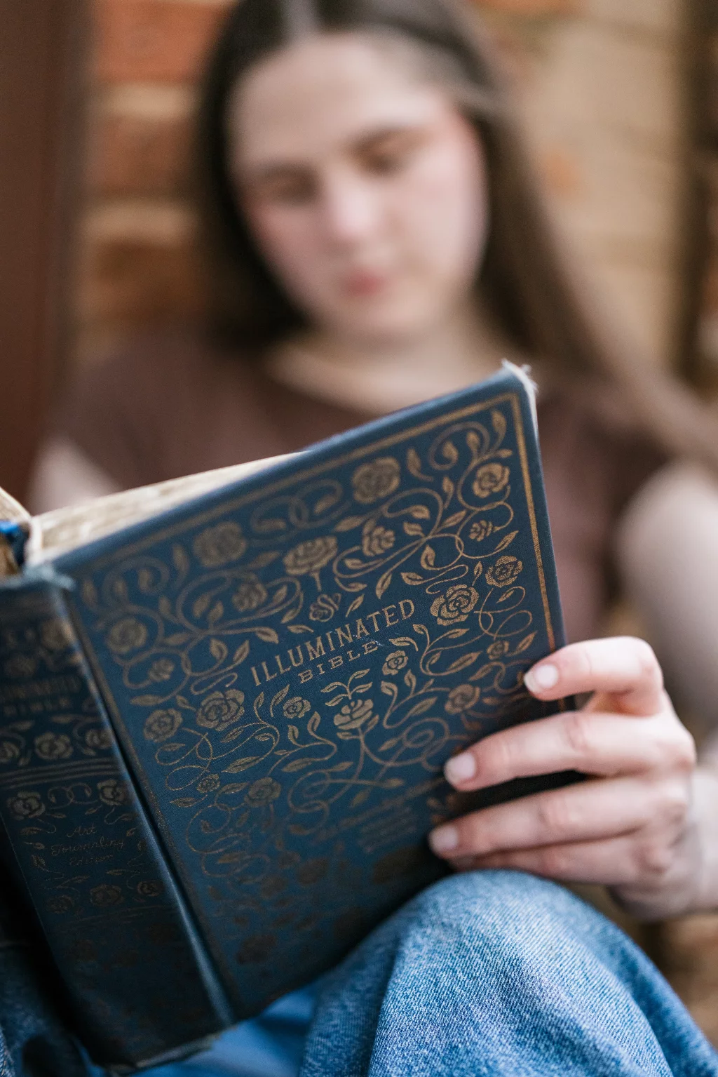 Kellis's senior photos taken Downtown, Greenville, SC: A person holding and reading a decorative book titled "Illuminated Bible" with gold floral designs on the cover. The person's face is out of focus in the background.