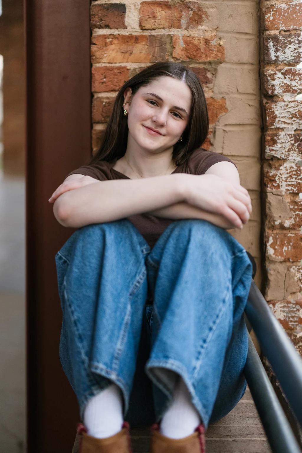 Kellis's senior photos taken Downtown, Greenville, SC: Young woman with long brown hair, wearing a brown shirt and blue jeans, sits on outdoor steps with her arms resting on her knees, smiling gently in front of a brick wall.