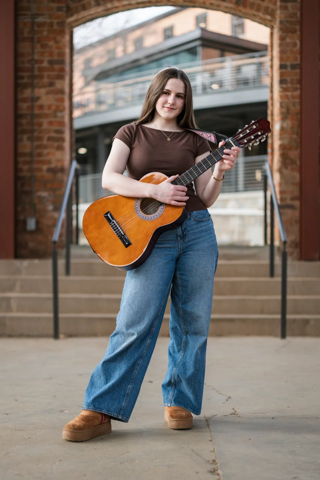 Kellis's senior photos taken Downtown, Greenville, SC: A young woman stands outdoors holding an acoustic guitar, wearing a brown shirt, blue jeans, and tan shoes, with a brick archway and building in the background.
