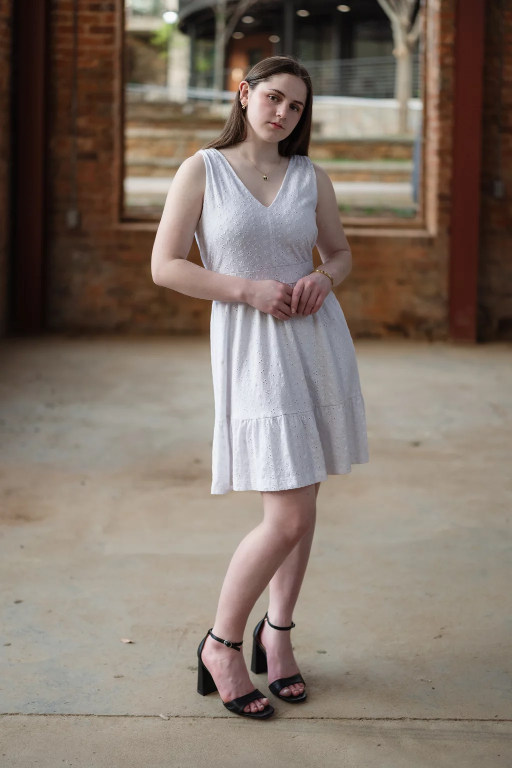 Kellis's senior photos taken Downtown, Greenville, SC: Young woman with straight brown hair wearing a sleeveless white dress and black high-heeled sandals, standing indoors on a concrete floor with exposed brick walls and large windows in the background.