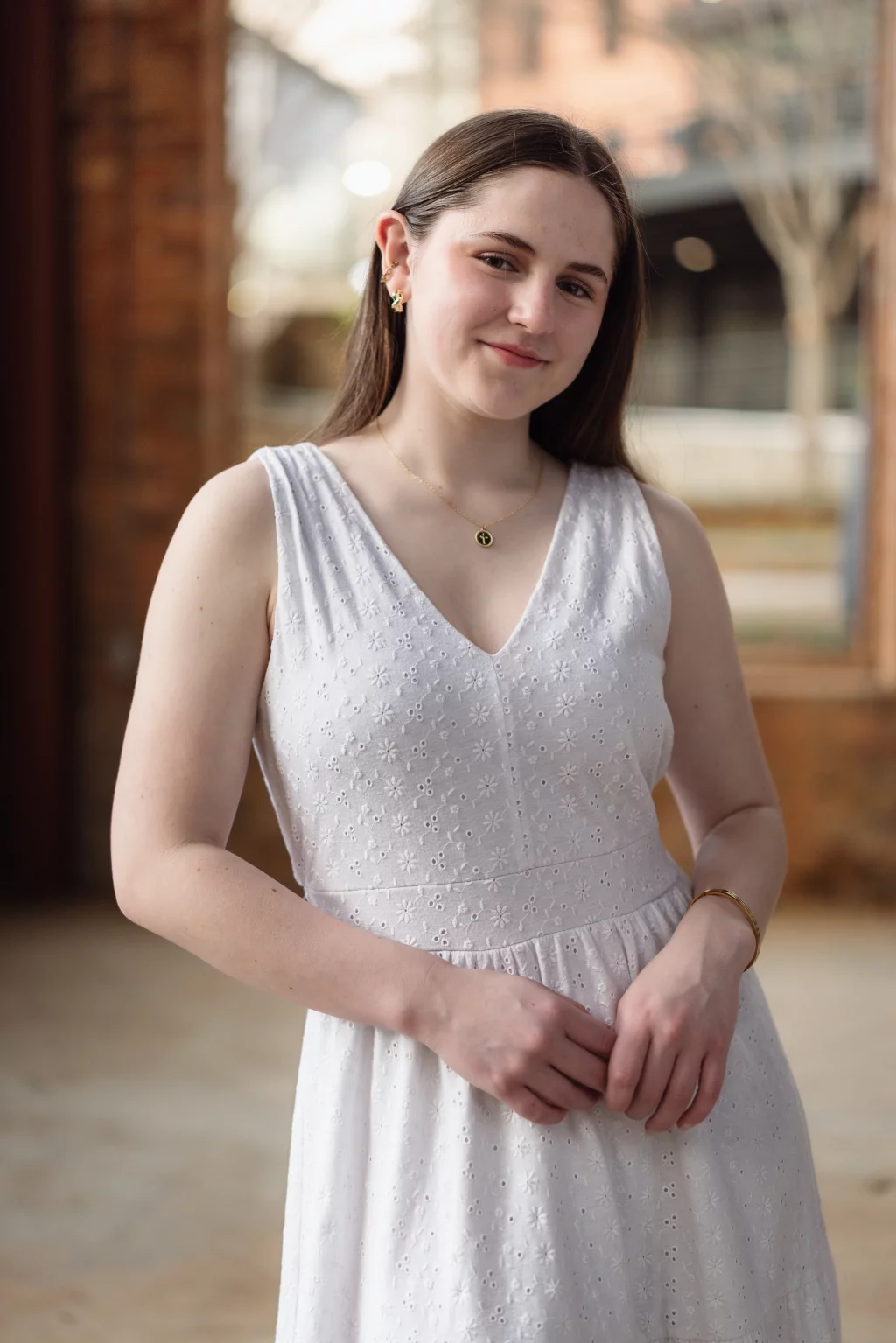 Kellis's senior photos taken Downtown, Greenville, SC: A young woman with long brown hair stands indoors, wearing a sleeveless white dress with a floral eyelet pattern, gold jewelry, and a subtle smile. The background is softly blurred with brick walls and large windows.