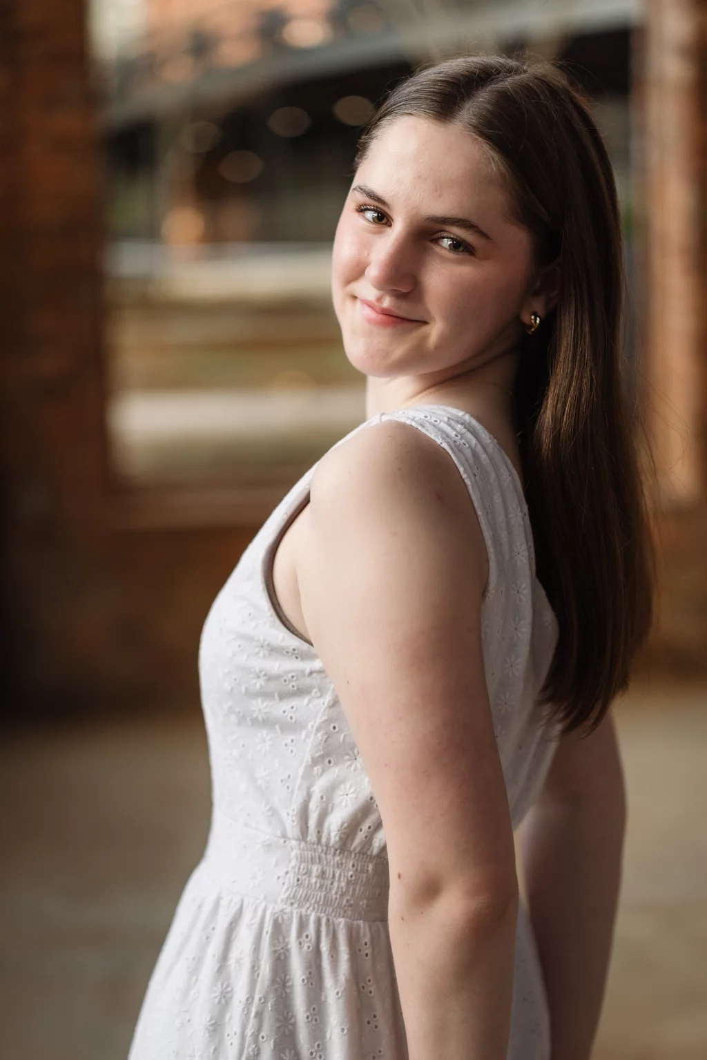 Kellis's senior photos taken Downtown, Greenville, SC: Young woman with long brown hair, wearing a sleeveless white dress, smiling and looking over her shoulder in a softly lit indoor setting with blurred background.