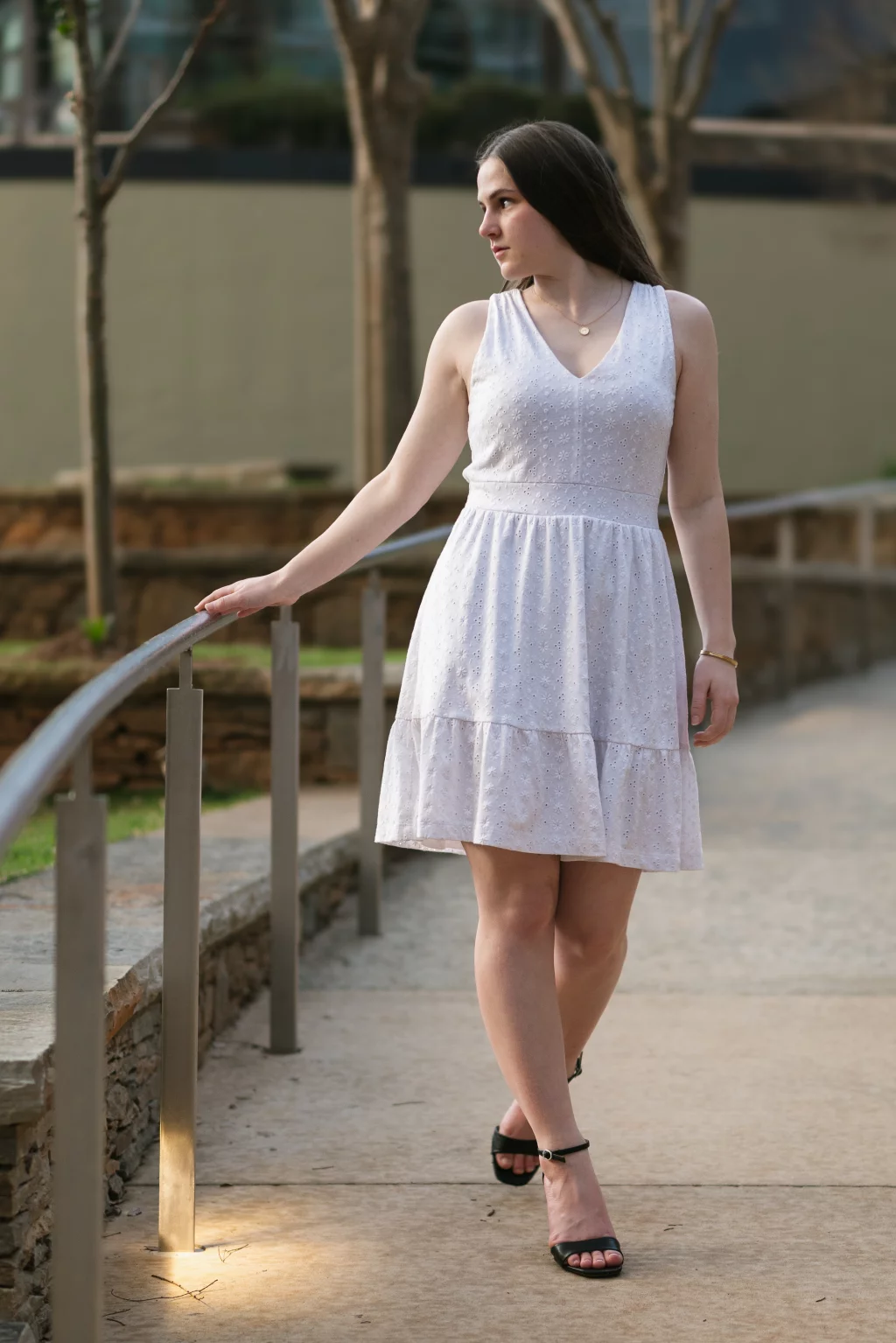 Kellis's senior photos taken Downtown, Greenville, SC: Woman in a sleeveless white dress and black sandals walking outdoors, holding onto a metal railing, with trees and a stone wall in the background.