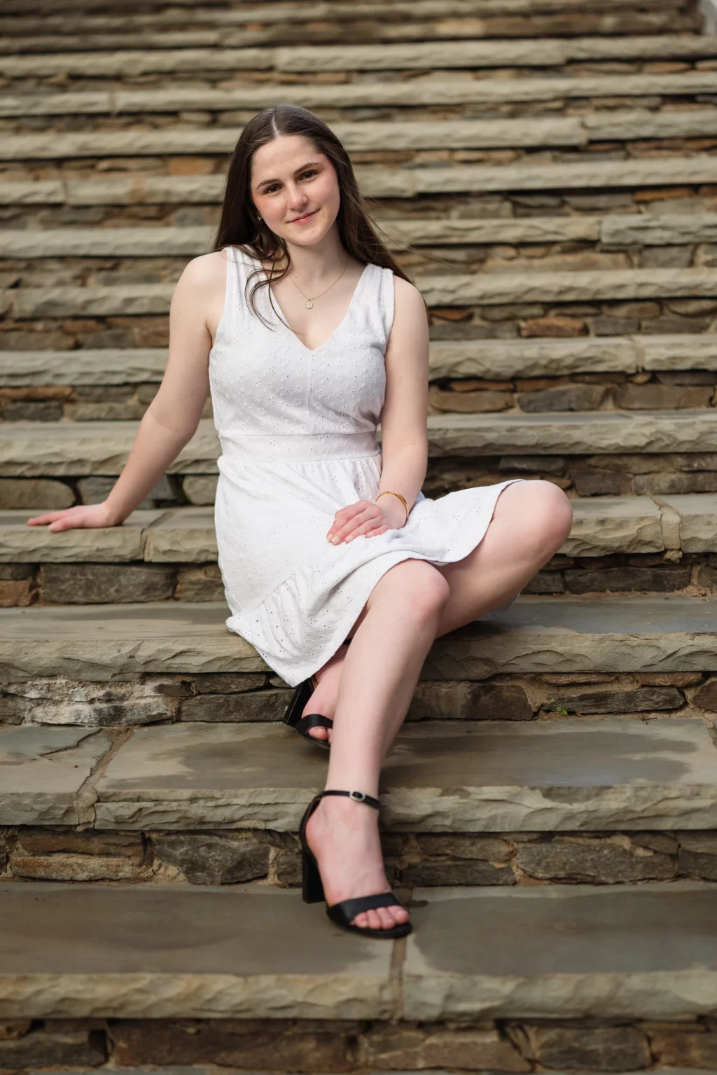 Kellis's senior photos taken Downtown, Greenville, SC: A young woman in a sleeveless white dress and black sandals sits on stone steps, smiling at the camera.