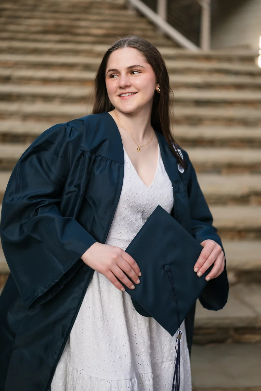 Kellis's senior photos taken Downtown, Greenville, SC: Young woman in a navy graduation gown and white dress holding a matching graduation cap, standing in front of stone steps and smiling to the side.