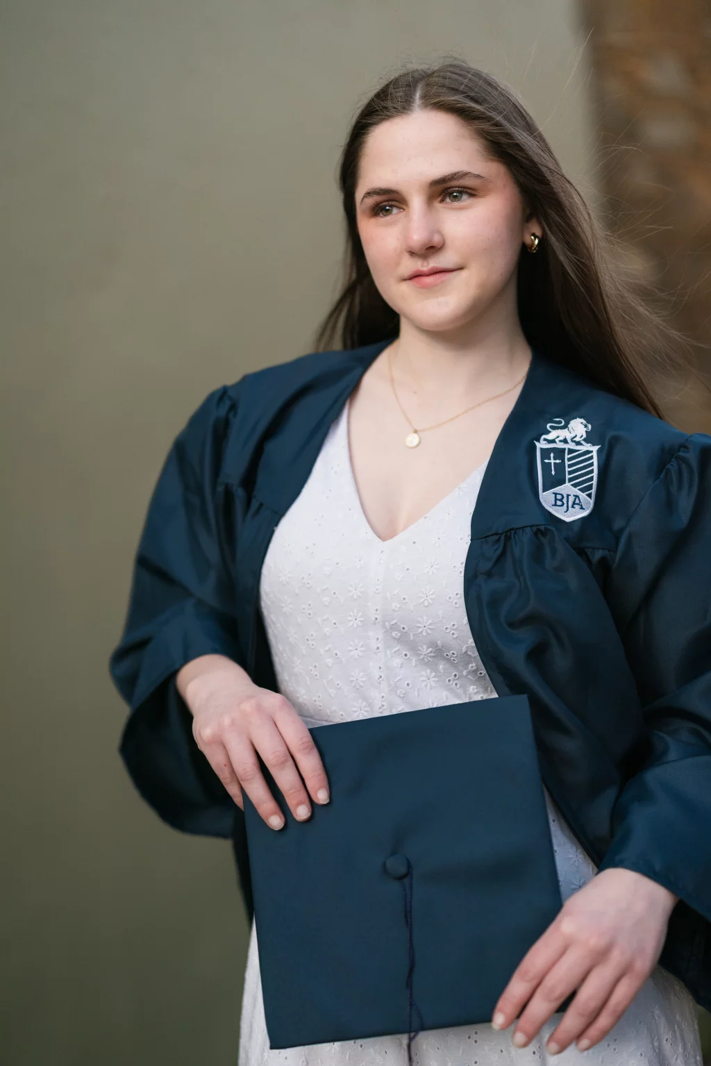 Kellis's senior photos taken Downtown, Greenville, SC: Young woman in a dark blue graduation gown holding a matching cap, with long brown hair and a neutral expression. She is wearing a white dress and a gold necklace. The gown has a crest with the letters "BJA.".