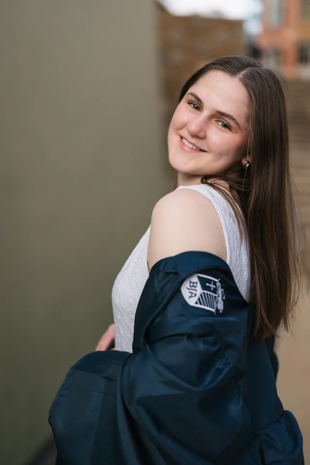 Kellis's senior photos taken Downtown, Greenville, SC: Young woman with long brown hair smiling over her shoulder, wearing a white sleeveless top and a dark jacket draped off one shoulder, with a patch on the sleeve.