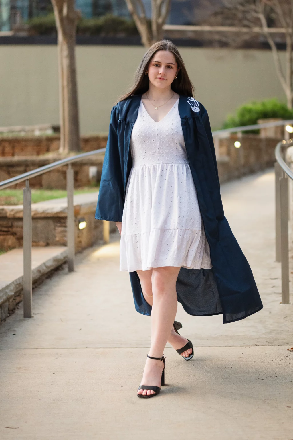 Kellis's senior photos taken Downtown, Greenville, SC: Young woman walking outdoors, wearing a white dress, dark graduation gown, and black heels, with long hair and a calm expression.