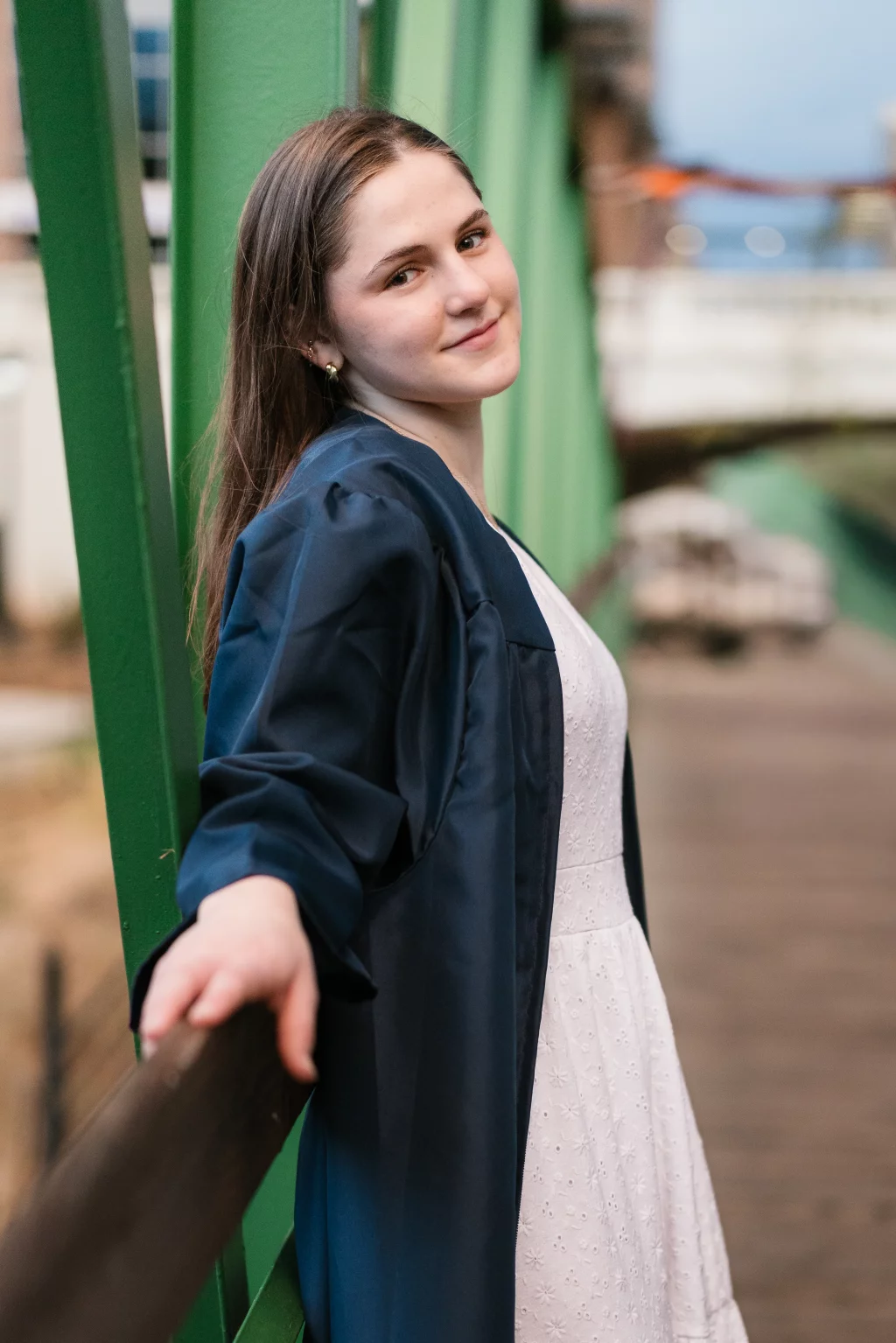 Kellis's senior photos taken Downtown, Greenville, SC: Young woman in a white dress and navy graduation gown leaning on a railing, smiling at the camera, standing on a green metal bridge.