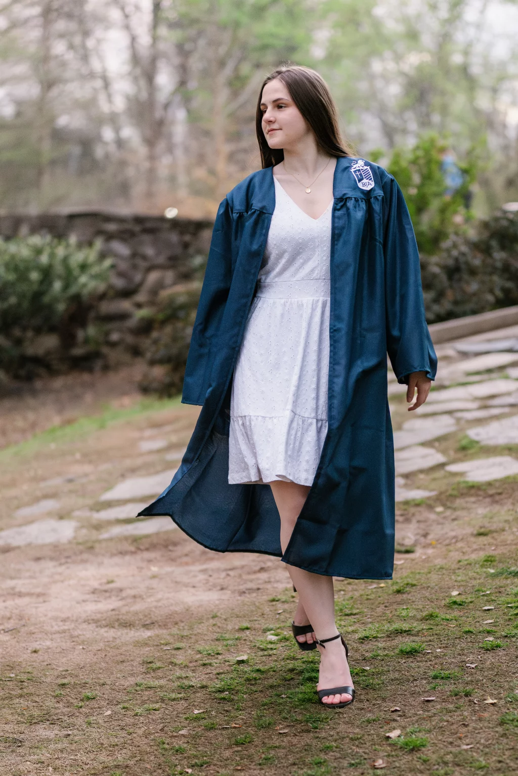 Kellis's senior photos taken Downtown, Greenville, SC: A young woman in a blue graduation gown over a white dress walks outdoors on a stone path, with greenery and a stone wall in the background.