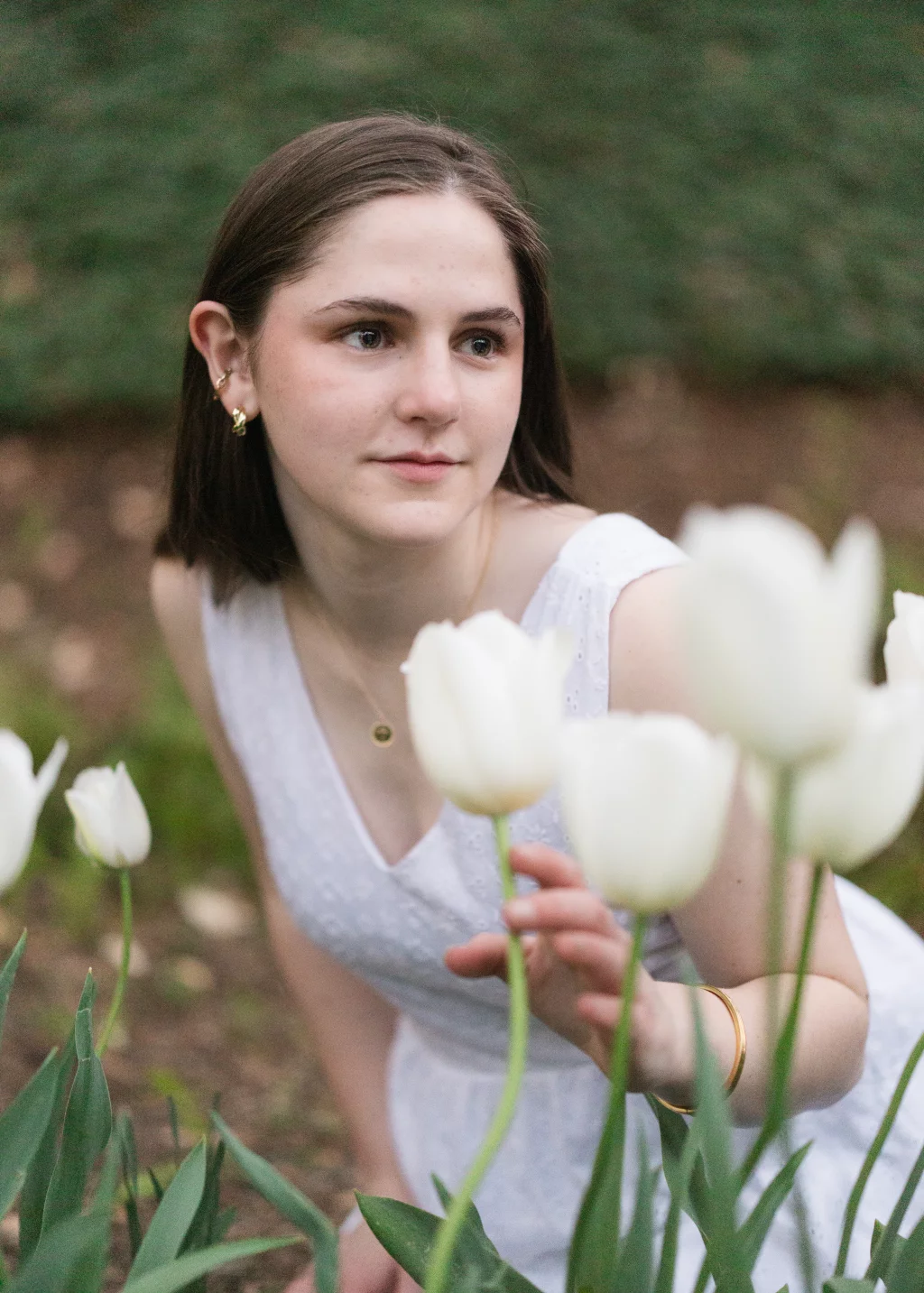 Kellis's senior photos taken Downtown, Greenville, SC: Young woman in a white dress crouching among white tulips, gently touching one flower, with a soft-focused green background.