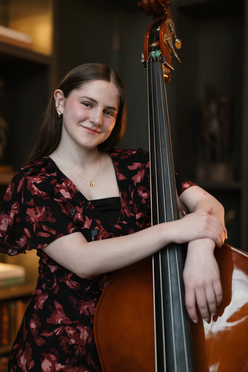Kellis's senior photos taken Downtown, Greenville, SC: Young woman with long brown hair, wearing a black and red floral dress, smiles and stands holding a double bass.