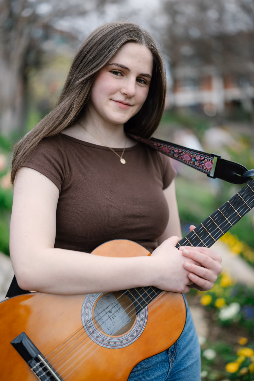 Kellis' senior photos taken Downtown Greenville, SC: Teen girl with long brown hair, wearing a brown shirt and blue jeans, standing outdoors holding an acoustic guitar with a patterned strap, with blurred greenery and flowers in the background.