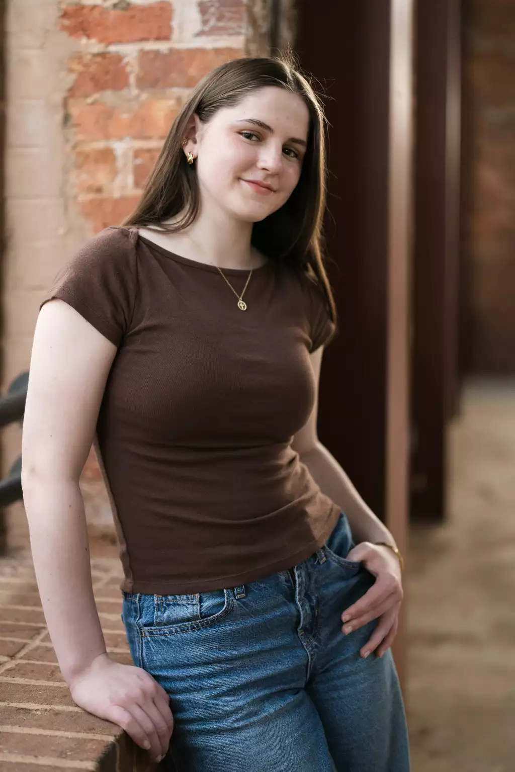 Kellis's senior photos taken Downtown, Greenville, SC: Young woman with long brown hair wearing a brown t-shirt and blue jeans, leaning against a brick wall and smiling slightly.