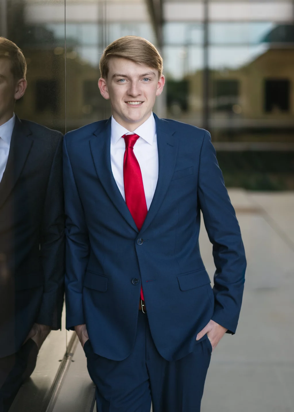 Macon's senior photos taken at Country Square and Downtown Greenville: Young man in a blue suit and red tie standing with one hand in his pocket, leaning against a reflective glass wall, smiling at the camera.