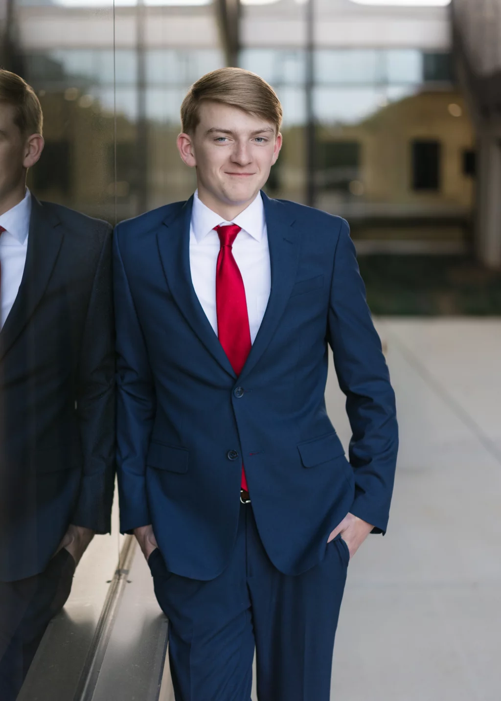 Macon's senior photos taken at Country Square and Downtown Greenville: Young man in a blue suit and red tie standing with hands in pockets, leaning against a reflective glass wall outdoors.