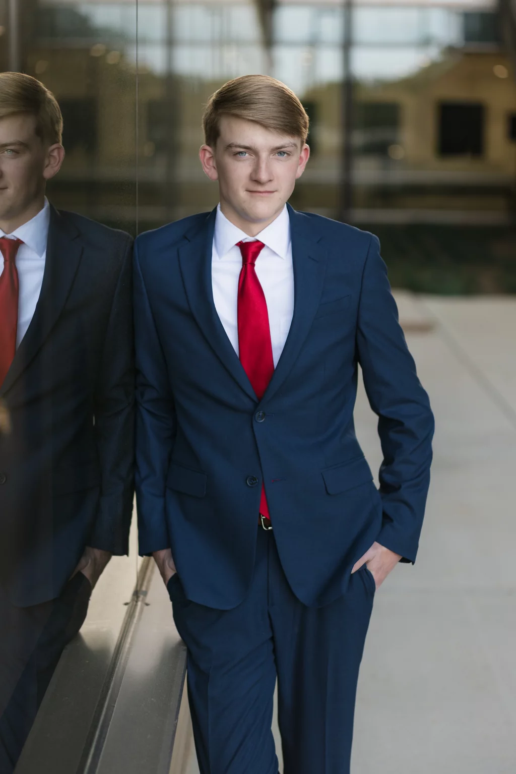Macon's senior photos taken at Country Square and Downtown Greenville: Young man in a blue suit with a red tie, standing with hands in pockets next to a reflective glass wall, looking at the camera.