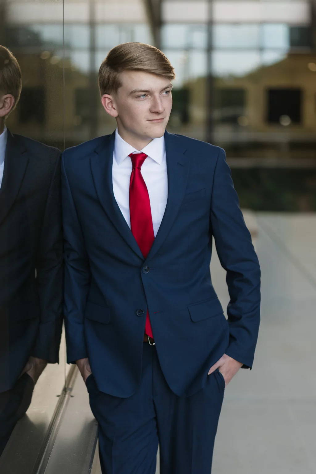 Macon's senior photos taken at Country Square and Downtown Greenville: Young man in a blue suit with a red tie standing with hands in pockets, leaning against a reflective glass wall, looking to the side.