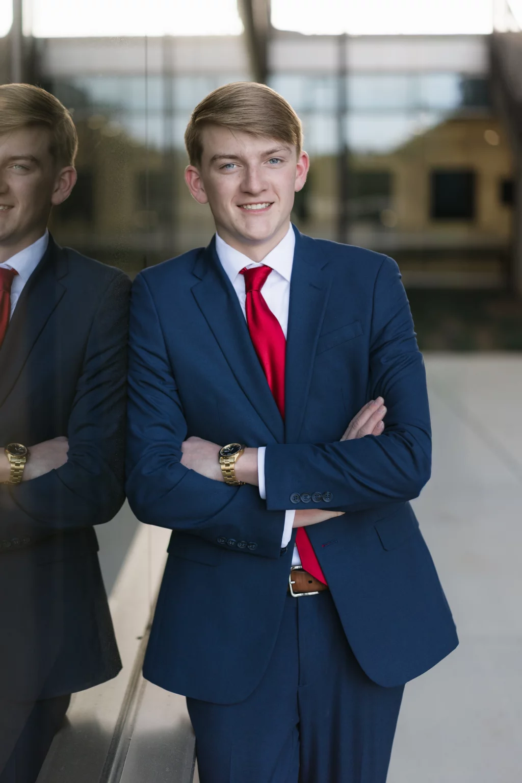 Macon's senior photos taken at Country Square and Downtown Greenville: Young man in a blue suit and red tie standing with arms crossed, smiling, and leaning against a glass wall with his reflection visible.