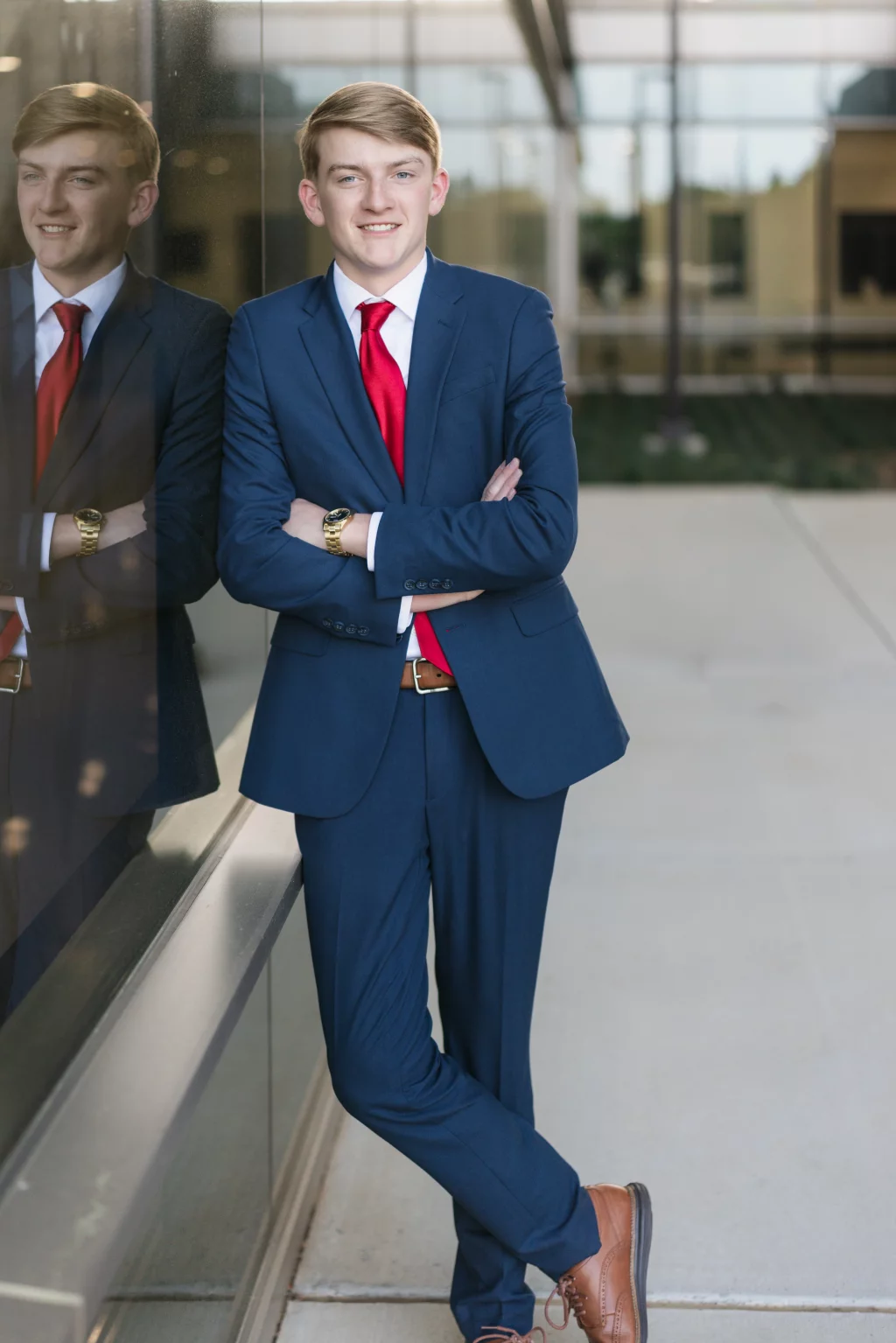 Macon's senior photos taken at Country Square and Downtown Greenville: Young man in a blue suit with a red tie and brown shoes, standing with arms crossed and leaning against a glass wall, his reflection visible beside him.