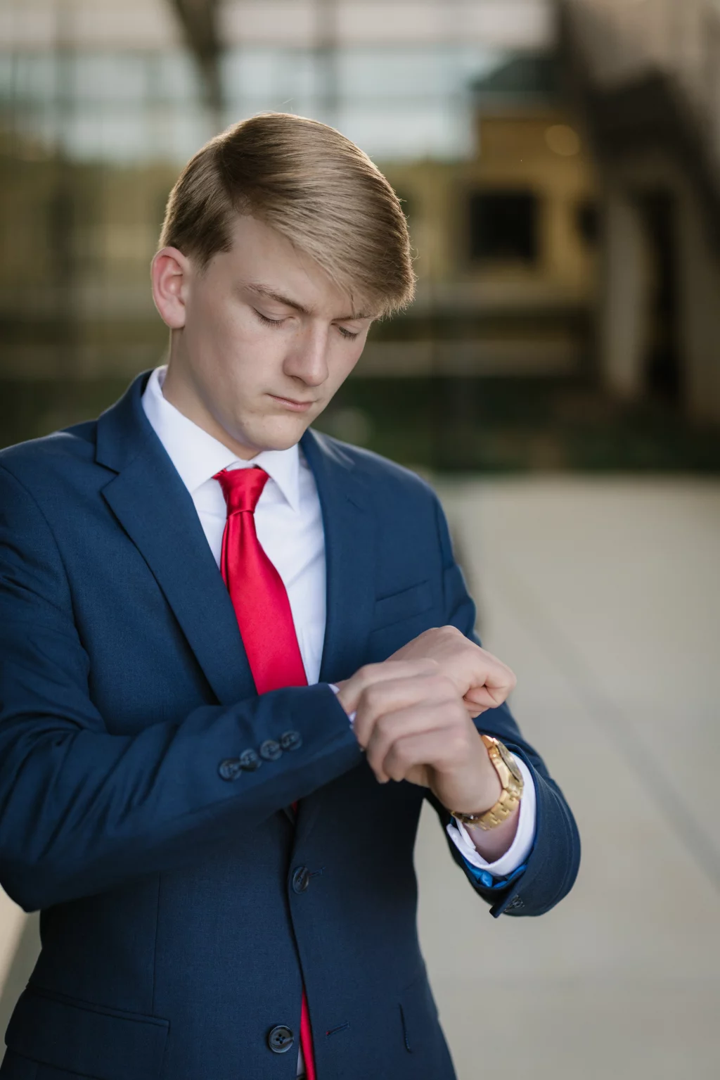 Macon's senior photos taken at Country Square and Downtown Greenville: Young man in a blue suit and red tie looking down at his wristwatch, standing outdoors with a blurred background.