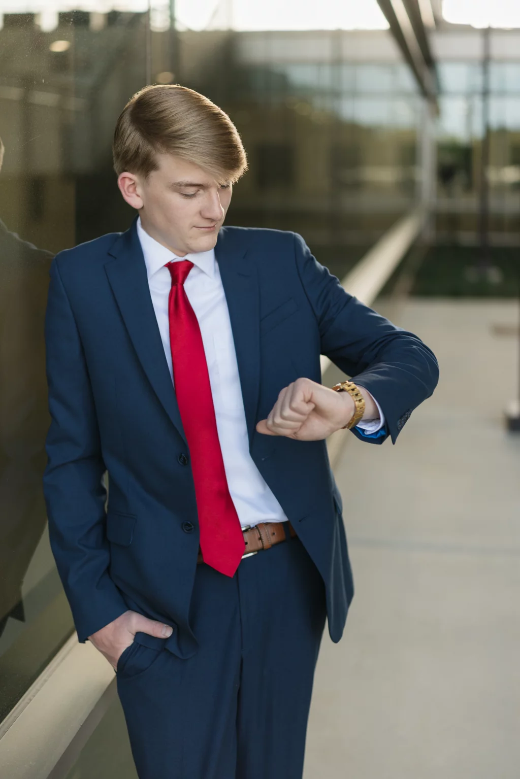 Macon's senior photos taken at Country Square and Downtown Greenville: Young man in a navy suit and red tie standing outdoors, looking at his watch with one hand in his pocket.