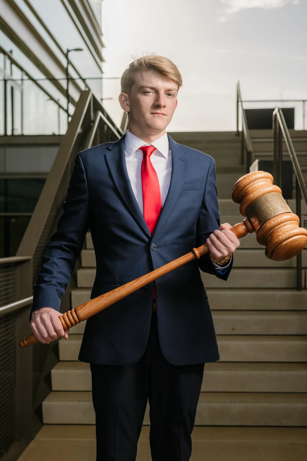 Macon's senior photos taken at Country Square and Downtown Greenville: Young man in a dark blue suit and red tie standing on stairs, holding an oversized wooden gavel.