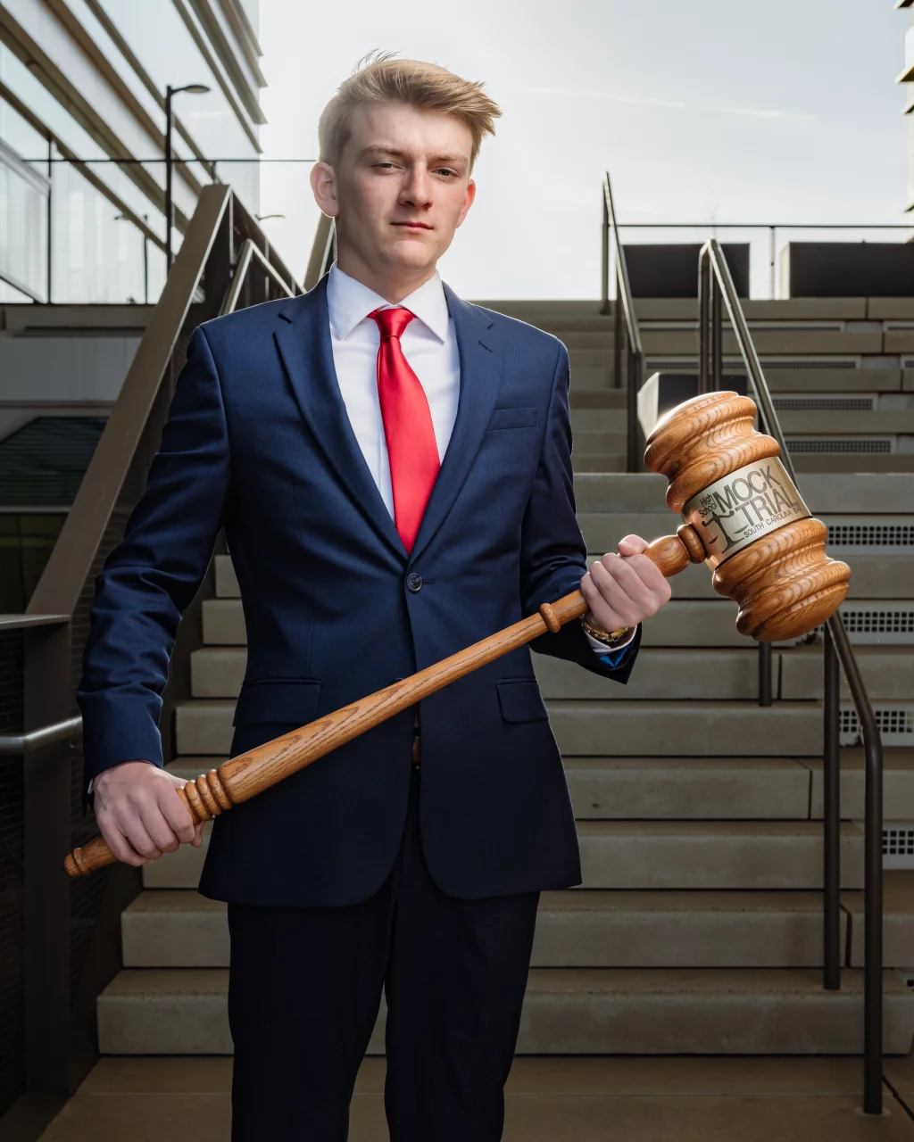 Macon's senior photos taken at Country Square and Downtown Greenville: Young man in a navy suit and red tie stands on steps outdoors, holding an oversized wooden gavel labeled "Mock Trial.".