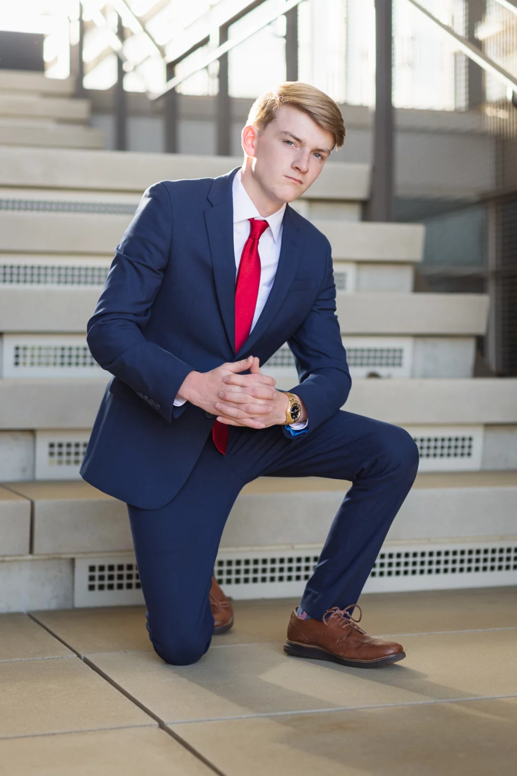 Macon's senior photos taken at Country Square and Downtown Greenville: Young man in a blue suit and red tie kneeling on one knee on outdoor steps, hands clasped in front, looking at the camera.