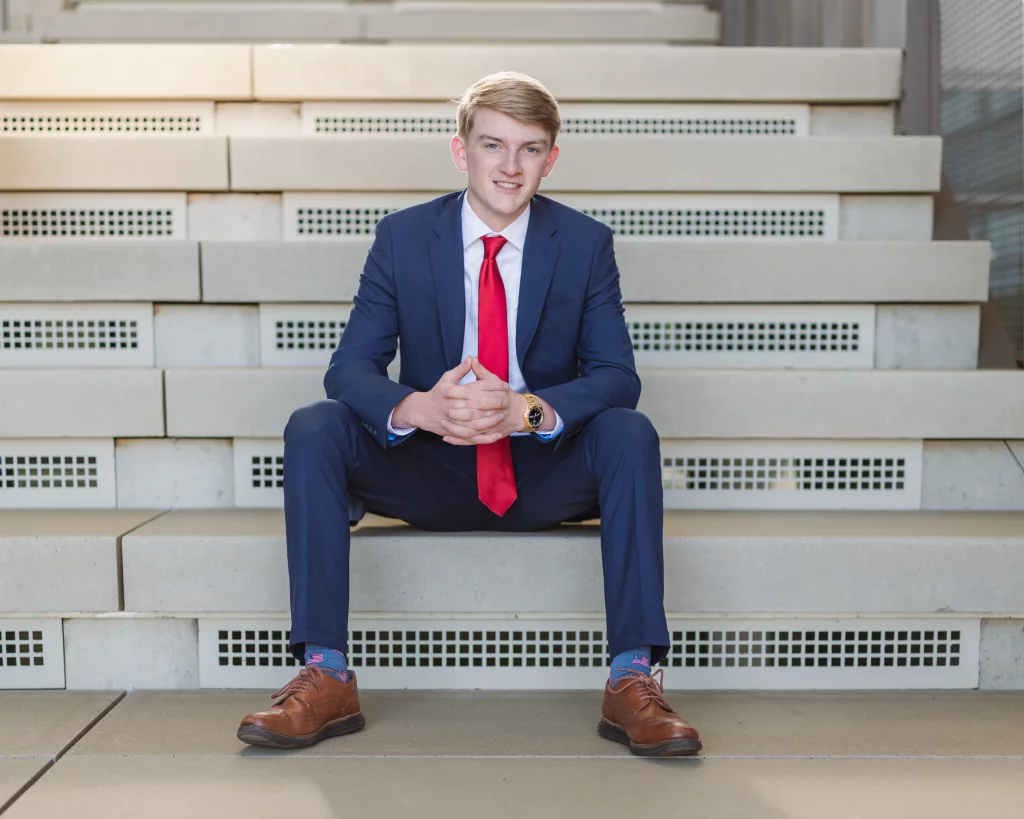 Macon's senior photos taken at Country Square and Downtown Greenville: Young man in a blue suit and red tie sitting on wide concrete steps, hands clasped, smiling at the camera.