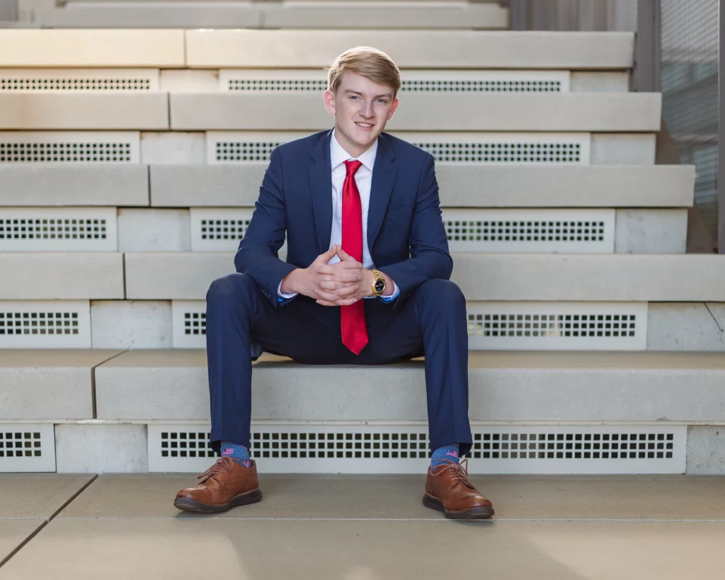 Macon's senior photos taken at Country Square and Downtown Greenville: Young man in a blue suit with a red tie and brown shoes sitting on concrete steps, hands clasped, looking at the camera and smiling.