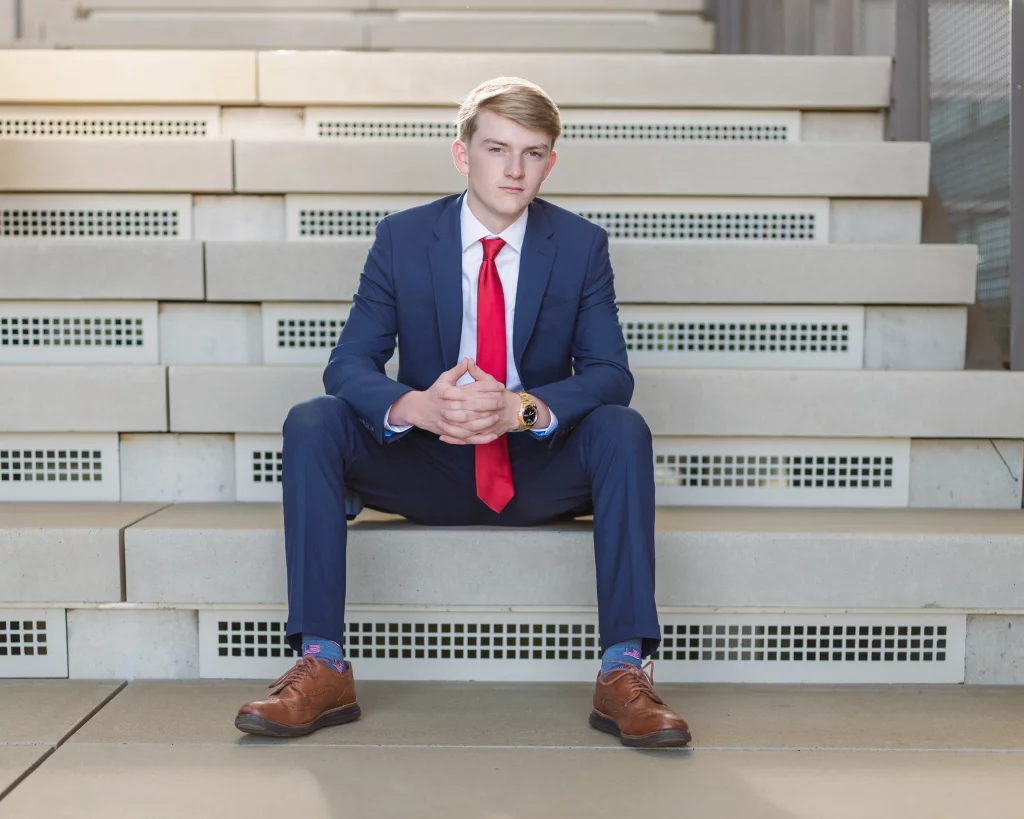 Macon's senior photos taken at Country Square and Downtown Greenville: Young man in a blue suit with a red tie and brown shoes sitting on concrete steps, looking directly at the camera with hands clasped.