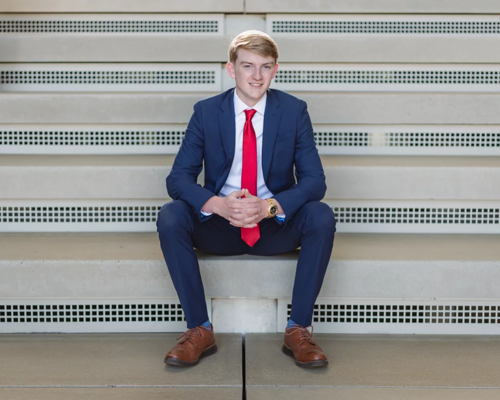 Macon's senior photos taken at Country Square and Downtown Greenville: Young man in a blue suit with a red tie and brown shoes, sitting on concrete steps, hands clasped, smiling at the camera.