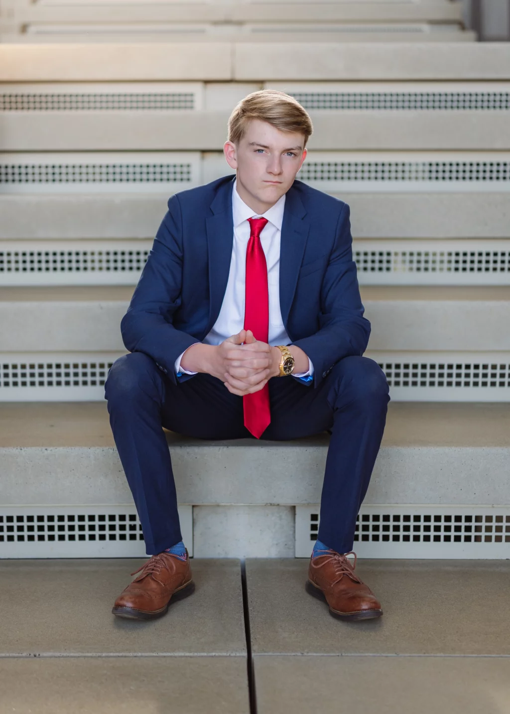 Macon's senior photos taken at Country Square and Downtown Greenville: Young man in a navy suit, white shirt, and red tie sitting on steps with hands clasped, looking serious.