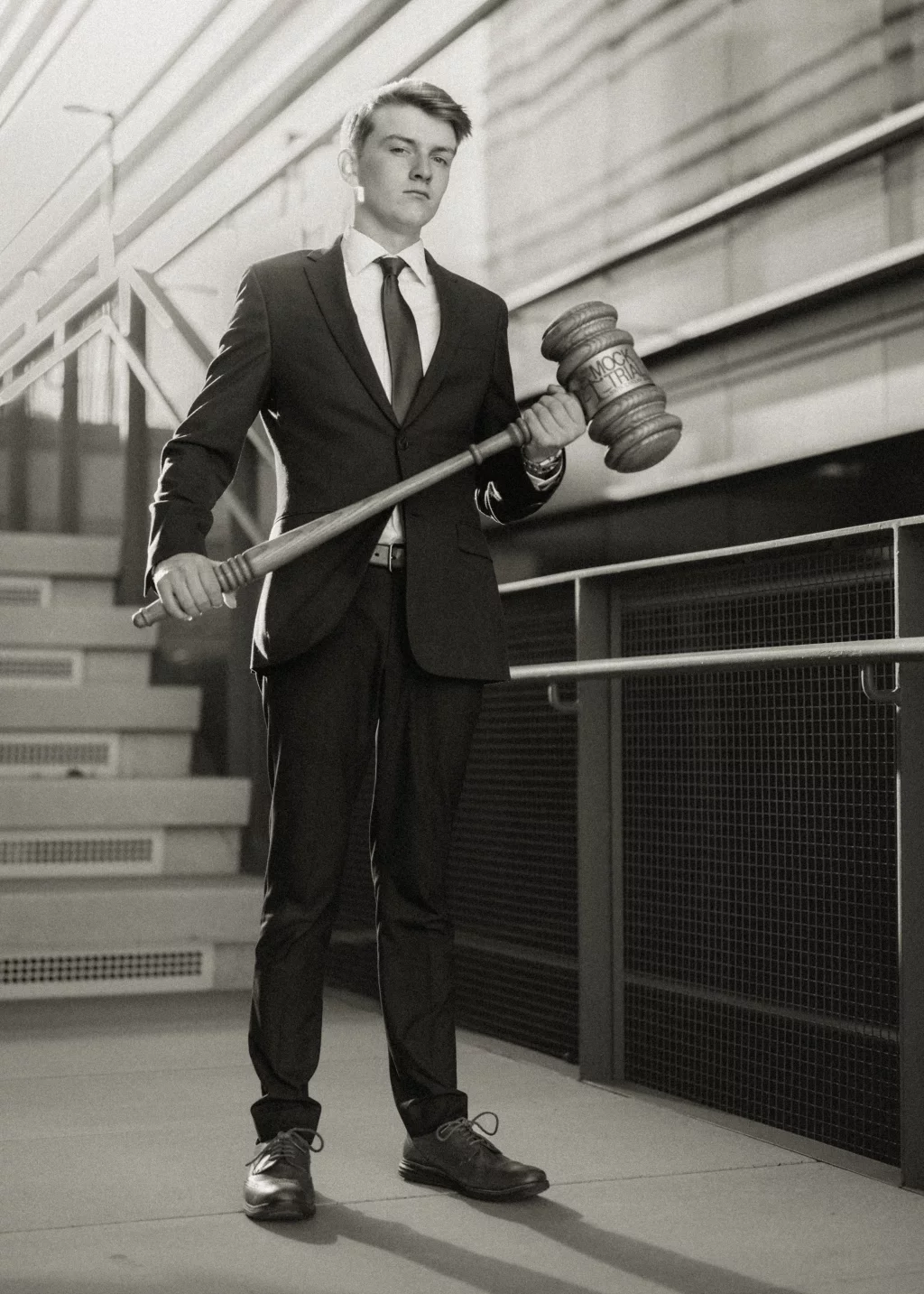 Macon's senior photos taken at Country Square and Downtown Greenville: A young man in a suit stands on a staircase holding an oversized gavel labeled "MOCK TRIAL." The image is in black and white.