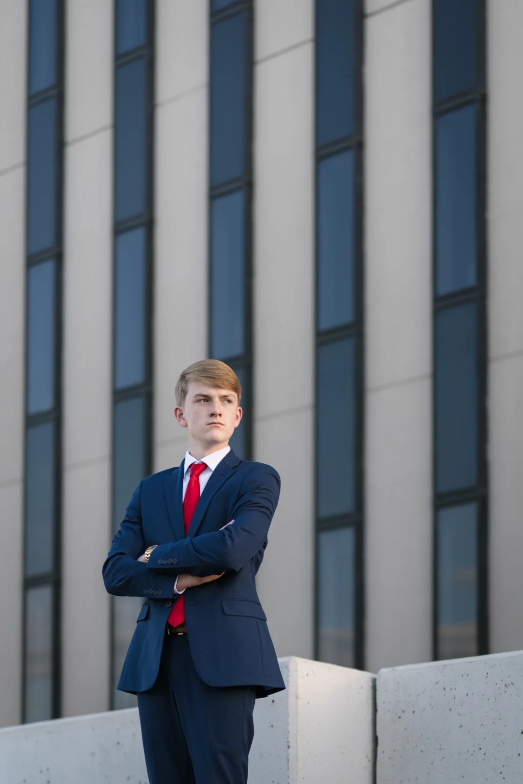 Macon's senior photos taken at Country Square and Downtown Greenville: Young man in a navy suit and red tie stands with arms crossed in front of a modern building with tall, vertical windows.